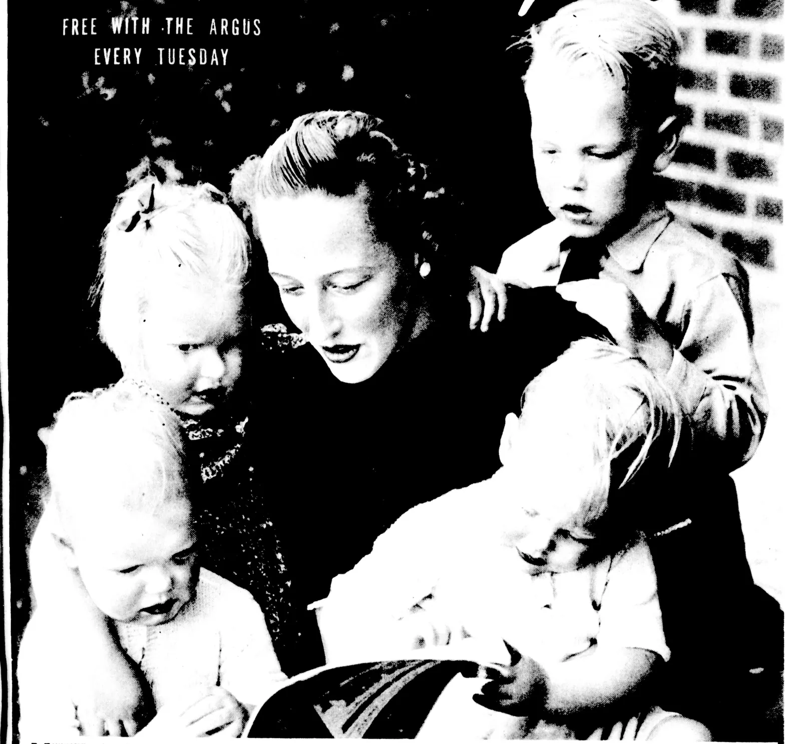 A newspaper photograph: a young woman with her hair swept up reads a book to four small fair-haired children crowded round her, two of them on her lap.