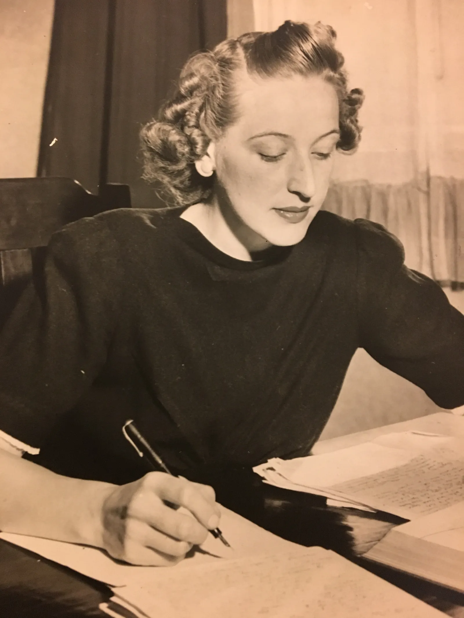 June Wright at a desk, writing with a fountain pen on a page of manuscript, eyes down, her hair in soft curls, in a dark high-necked dress; loose pages at her elbow and a curtained window behind.